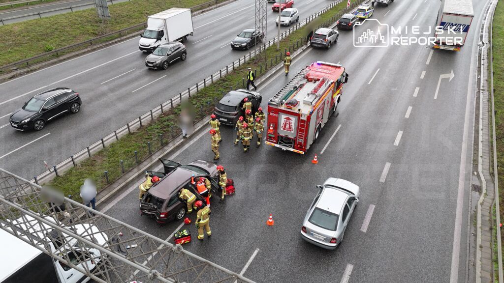 Grupa strażaków w hełmach pracuje przy uszkodzonym samochodzie na pasie ruchu, obok stoi wóz strażacki.