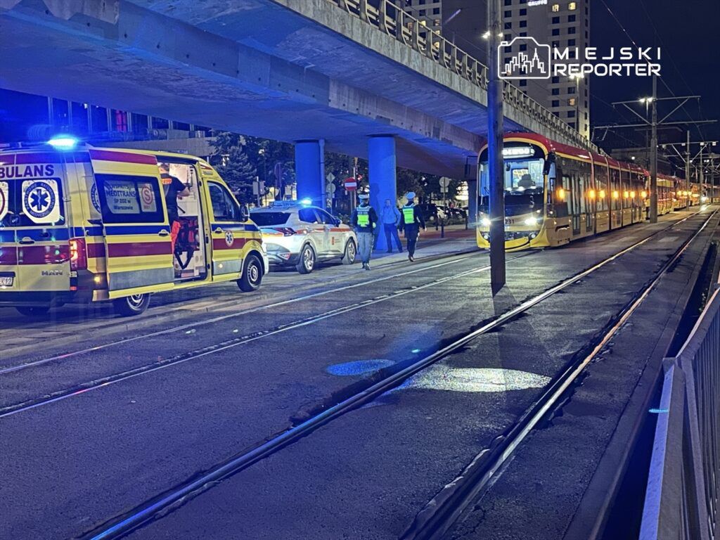 Karetka pogotowia oraz radiowóz stoją na ulicy, obok tramwaju, w nocy, w otoczeniu miejskim.