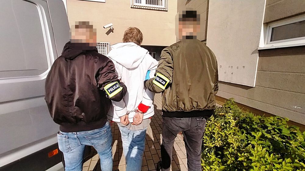 Mężczyzna w kajdankach prowadzony przez dwóch policjantów na zewnątrz budynku mieszkalnego.
