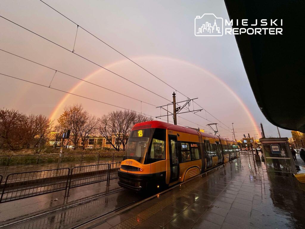 Tramwaj linii 18 zatrzymany na przystanku, w tle widoczna tęcza na tle szarego nieba i nagrzanych drzew.