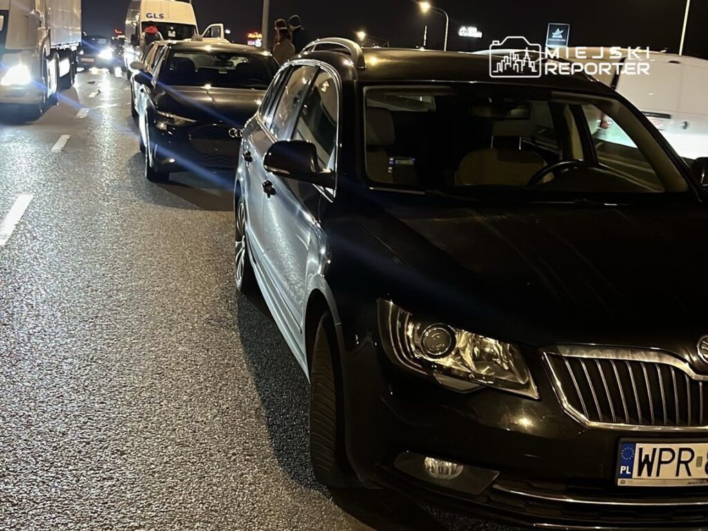 Zablokowana droga nocą z kilkoma samochodami stojącymi w korku, w tym czarną limuzyną i białym SUV-em.