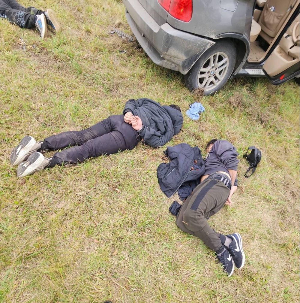 Dwie osoby leżą na trawie obok samochodu, jedna z nich ma ręce związane, obie w ciemnych ubraniach.