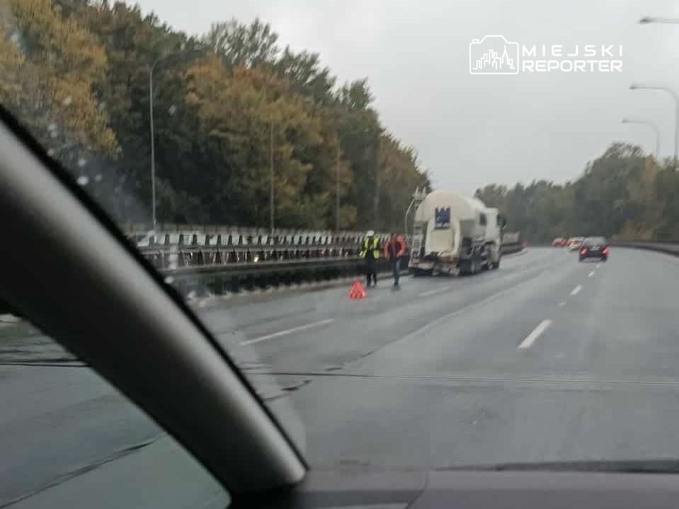 Kierowcy zatrzymują się obok uszkodzonej betoniarki na drodze, a pracownicy w kamizelkach odblaskowych zabezpieczają miejsce zdarzenia.