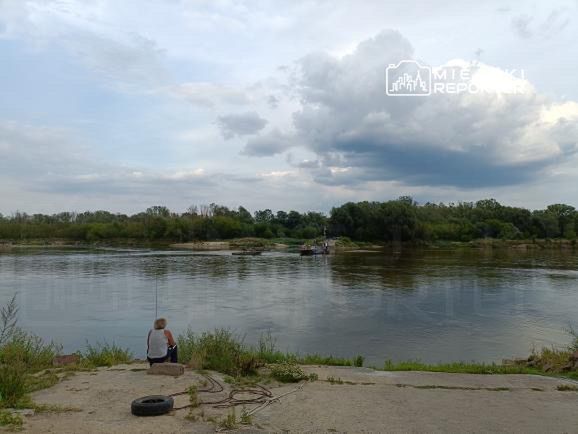 Mężczyzna siedzący na brzegu rzeki, wpatrujący się w wodę, z chmurami na niebie w tle.