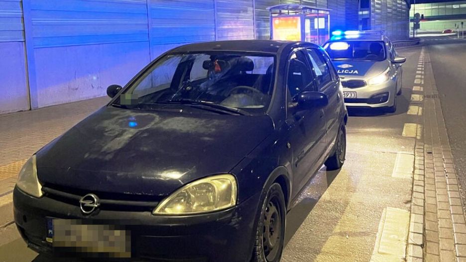 Czarny samochód osobowy zatrzymany na drodze nocą, obok policyjny radiowóz z włączonymi sygnałami świetlnymi.