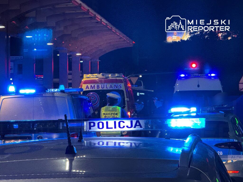 Grupa policjantów i ratowników medycznych pracuje przy ambulansie pod oświetlonym dworcem, otoczona pojazdami służb.