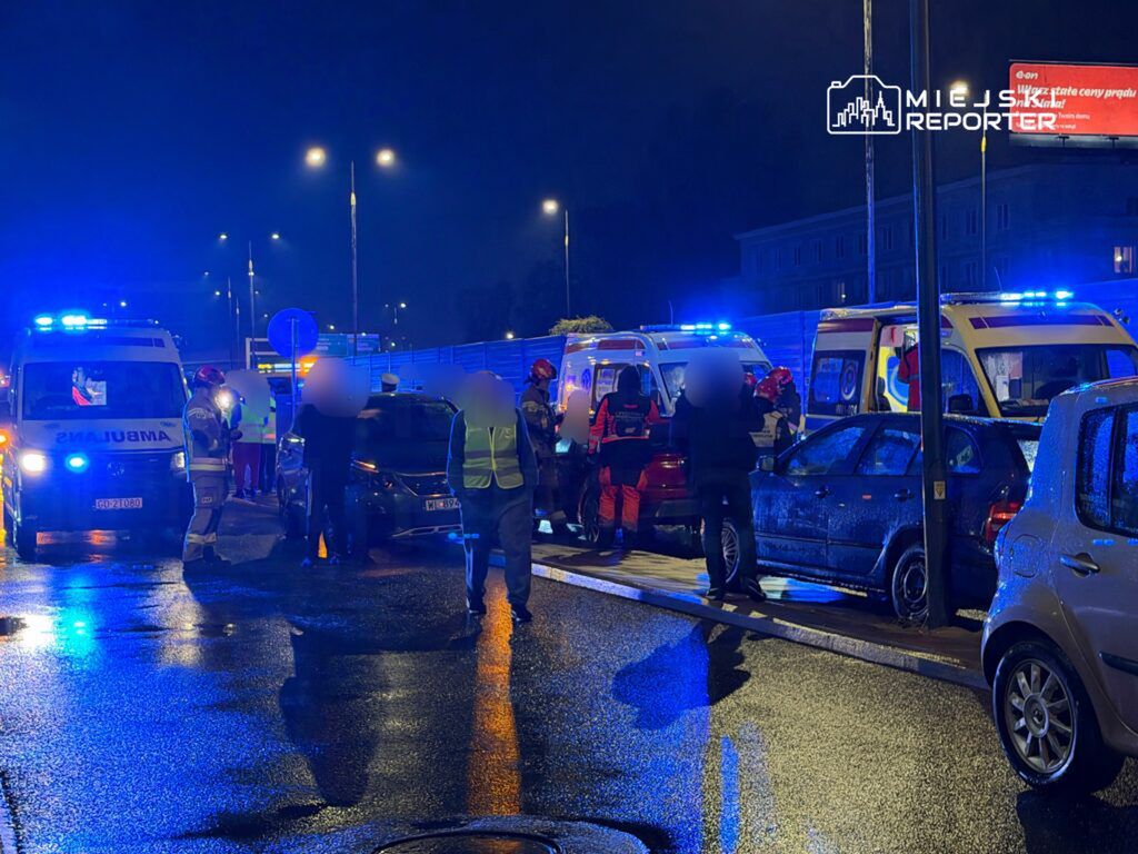 Grupa ratowników medycznych i policjantów udziela pomocy poszkodowanym po wypadku drogowym na mokrej jezdni w nocy.