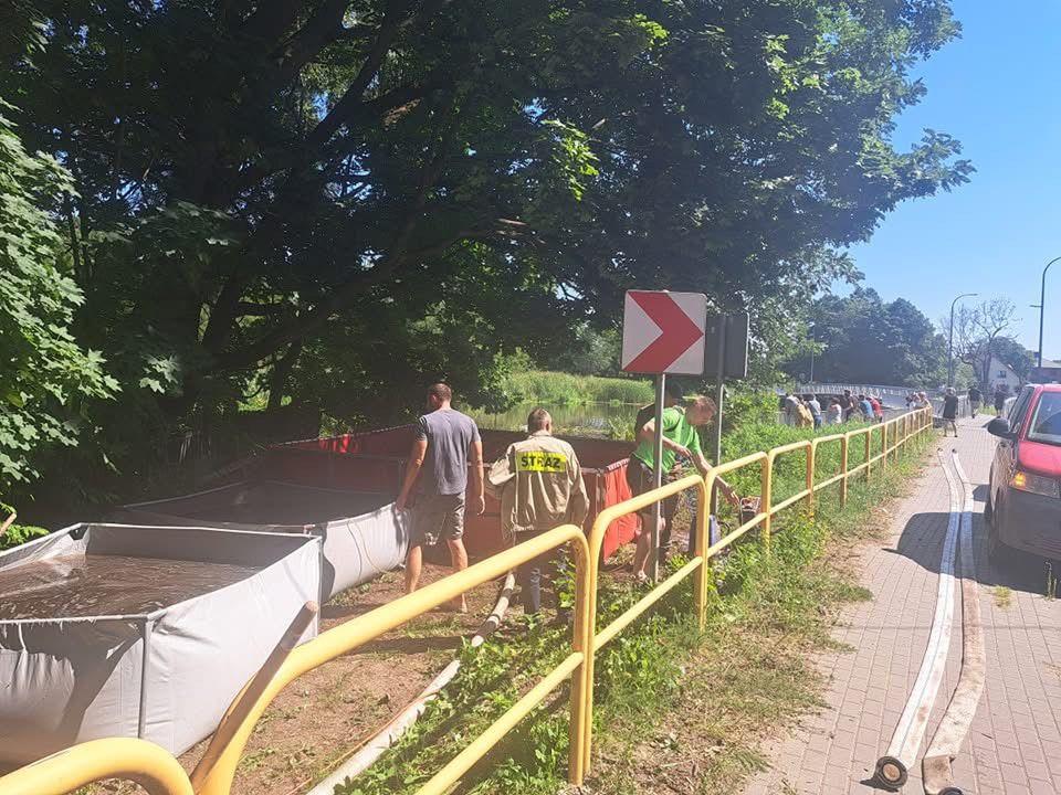 Grupa osób napełnia worki z piaskiem wzdłuż rzeki, obok żółtej bariery ochronnej, w słoneczny dzień.