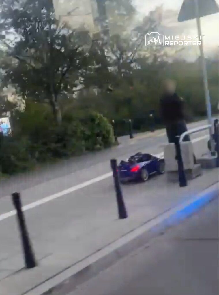 Dziecko w małym, niebieskim samochodziku zabawkowym porusza się po chodniku w parku.