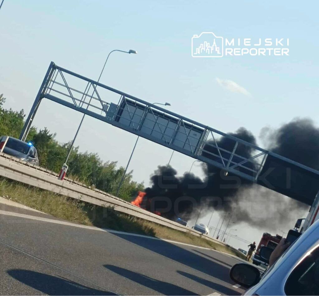 Pożar pojazdu na poboczu drogi, z dymem unoszącym się w powietrzu, obok policyjny radiowóz.