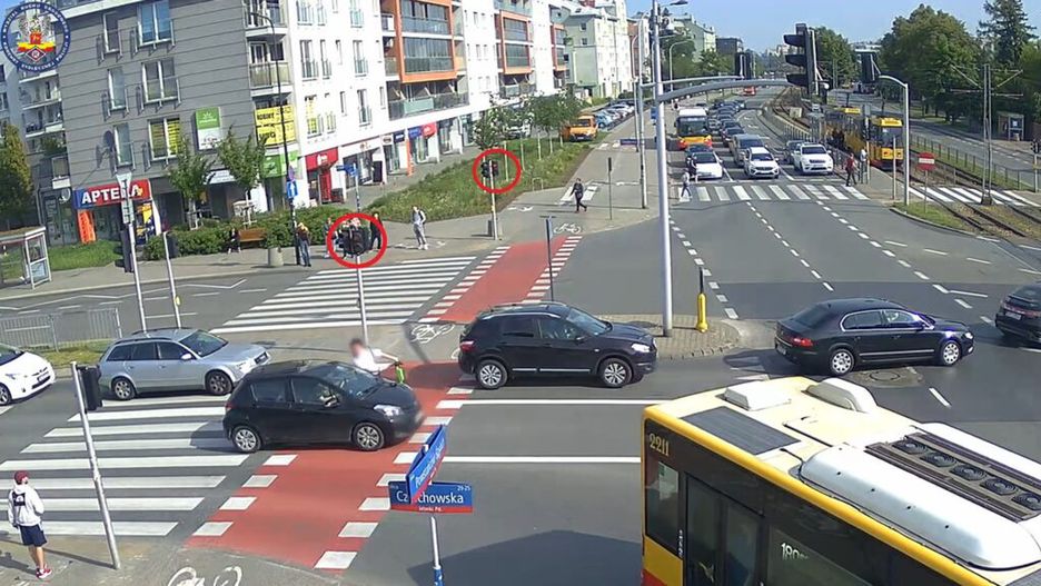 Grupa osób stojąca na chodniku przy przejściu dla pieszych, obok ruchliwej ulicy, w tle samochody i tramwaj.