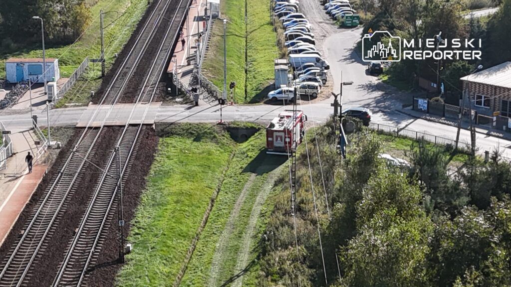 W tle torów kolejowych stoi wóz strażacki obok przejazdu, a w oddali widać zaparkowane samochody.