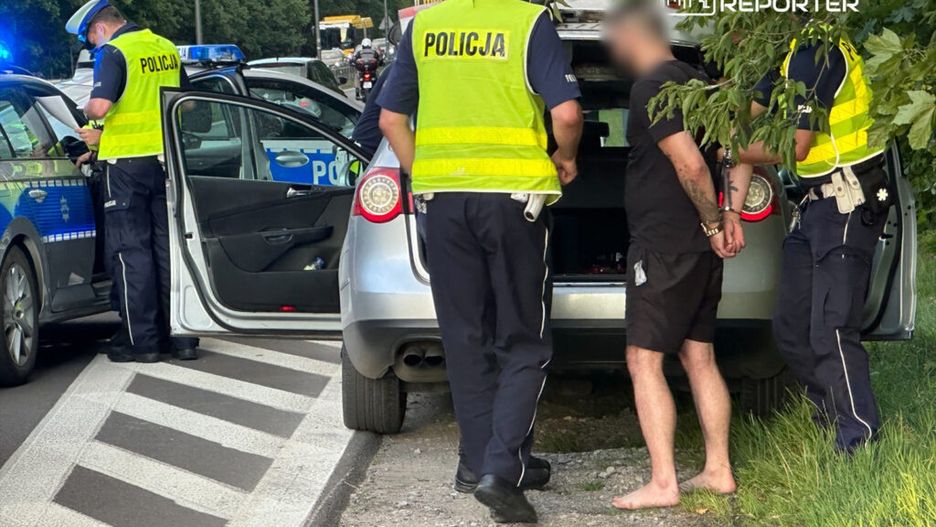 Mężczyzna w czarnej koszulce i szortach z rękami skrępowanymi przez policję stoi obok otwartego samochodu. Policjanci w kamizelkach odblaskowych sprawdzają dokumenty.