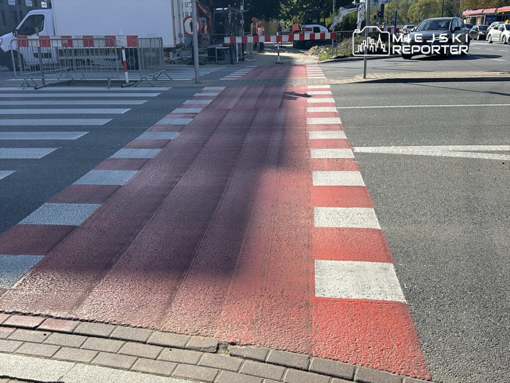 Pasy ruchu z czerwonymi i białymi oznaczeniami na przejściu dla pieszych w miejskim otoczeniu. W tle widoczne bariery budowlane.