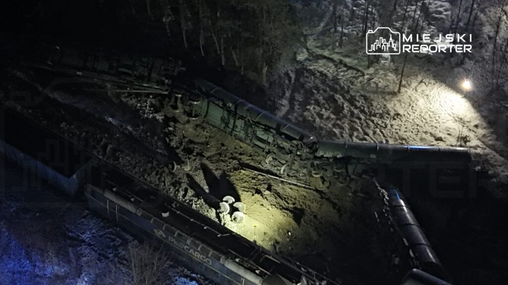 W nocy, w zalesionym terenie, wykolejony pociąg towarowy leży na boku w błocie, otoczony przez śnieg.