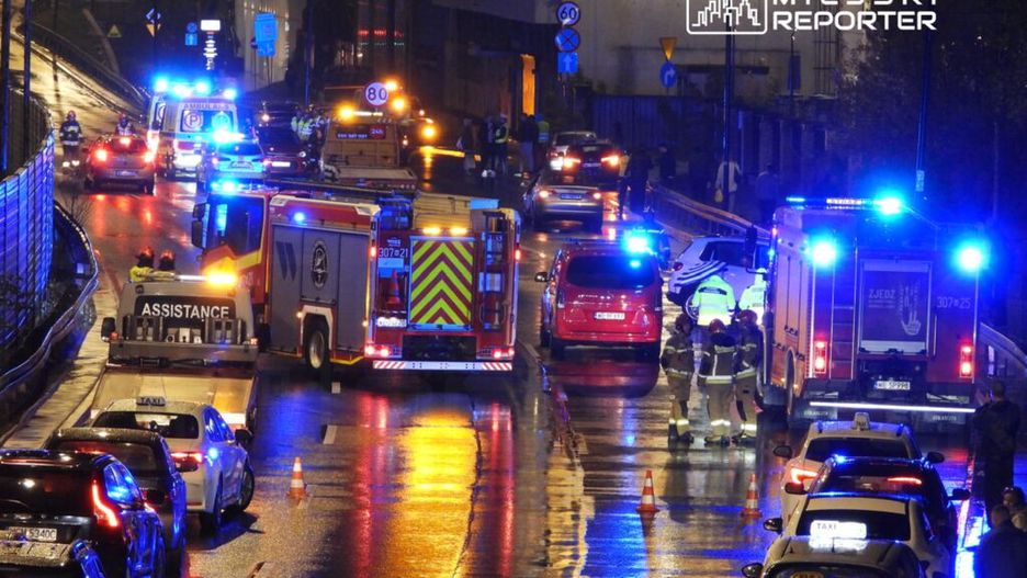 Na mokrej jezdni, w otoczeniu pojazdów ratunkowych, strażacy i ratownicy medyczni udzielają pomocy poszkodowanym.