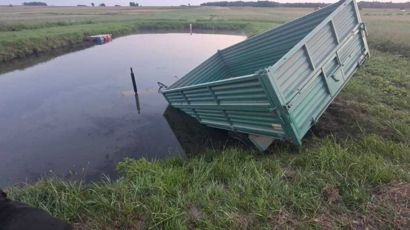 Zielona przyczepa leży na boku w wodzie, otoczona trawą i polami. W tle widoczne niskie wzgórza.