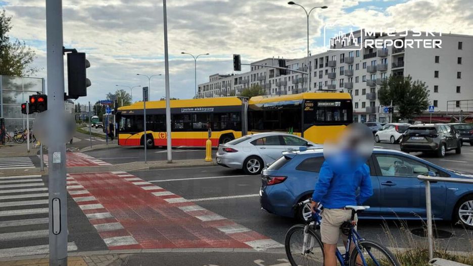 Autobus elektryczny skręca w lewo na ruchliwym skrzyżowaniu, a rowerzysta czeka przy krawężniku.