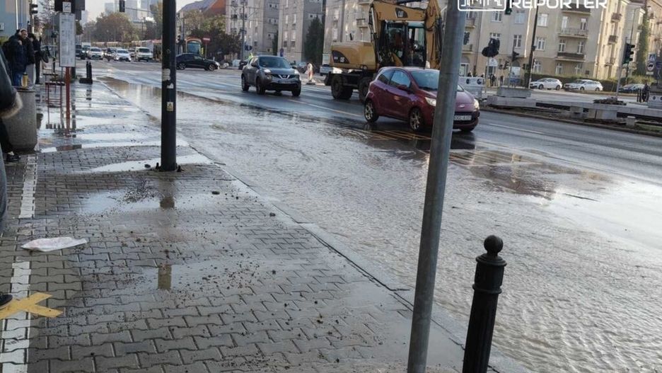 Ruchliwa ulica z samochodami, na której widoczne są kałuże wody i kontener wywożący ziemię, w tle budynki mieszkalne.