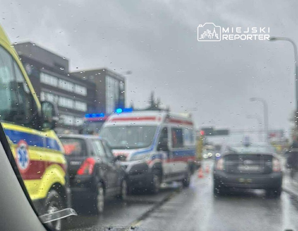 Ambulans z włączonymi sygnałami świetlnymi stoi na ruchliwej ulicy w deszczową pogodę, otoczony przez inne pojazdy.