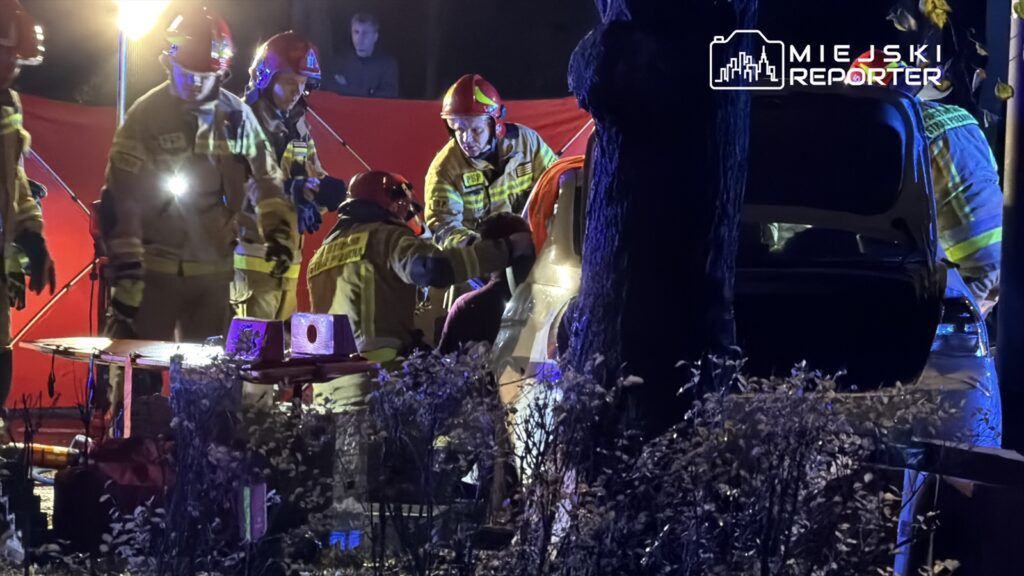 Grupa strażaków w mundurach ratunkowych pracuje przy samochodzie w nocy, otoczona jasnymi światłami i czerwonym tłem.