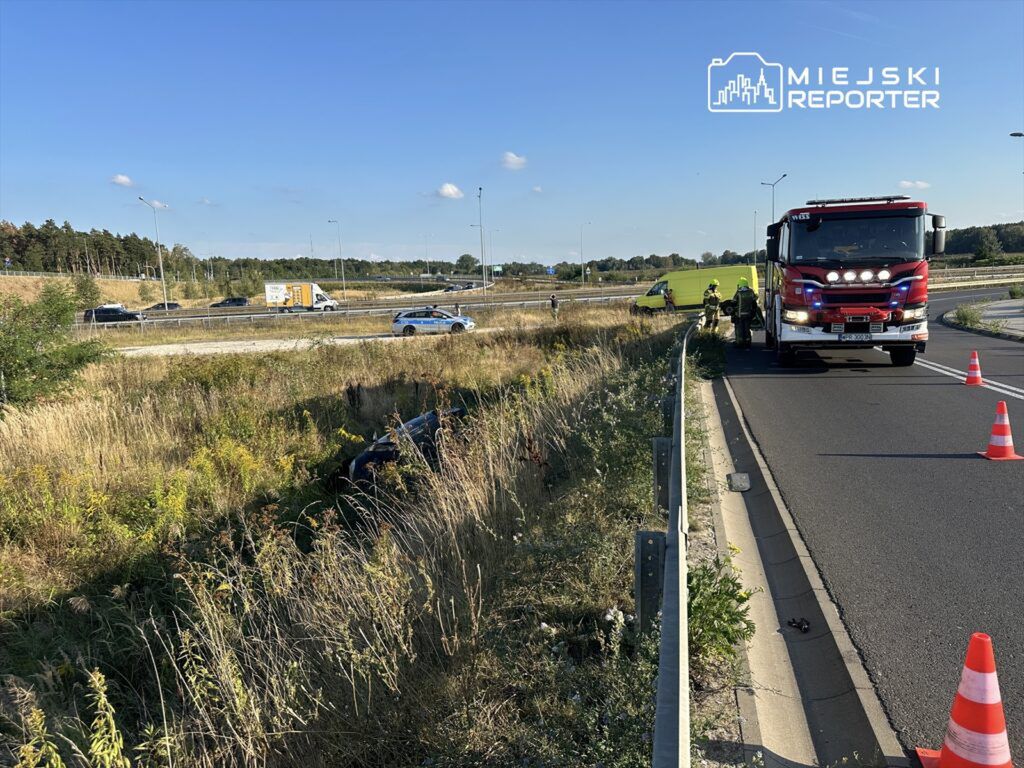 Na poboczu drogi stoi wóz strażacki, a obok pracują strażacy, zabezpieczając miejsce wypadku.