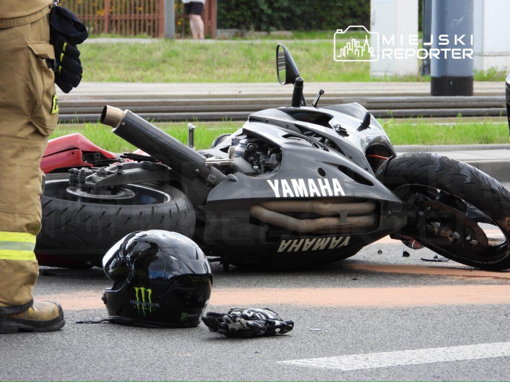 Motocykl marki Yamaha leży na boku na jezdni, obok niego znajduje się kask z logo Monster. W tle widoczny strażak w mundurze.