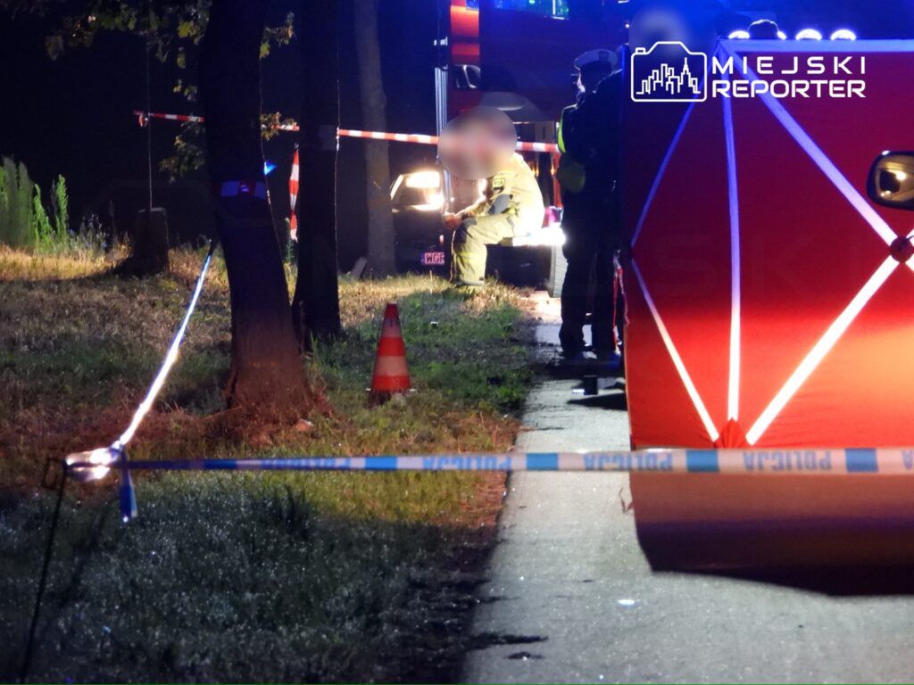 Na zdjęciu widać miejsce zdarzenia, gdzie interweniują służby ratunkowe. W tle znajduje się pojazd oraz oznakowanie policyjne. Funkcjonariusze zabezpieczają teren, a w pobliżu widać osobę w mundurze, która prowadzi działania. Scena miała miejsce w nocy, co podkreśla oświetlenie służb.
