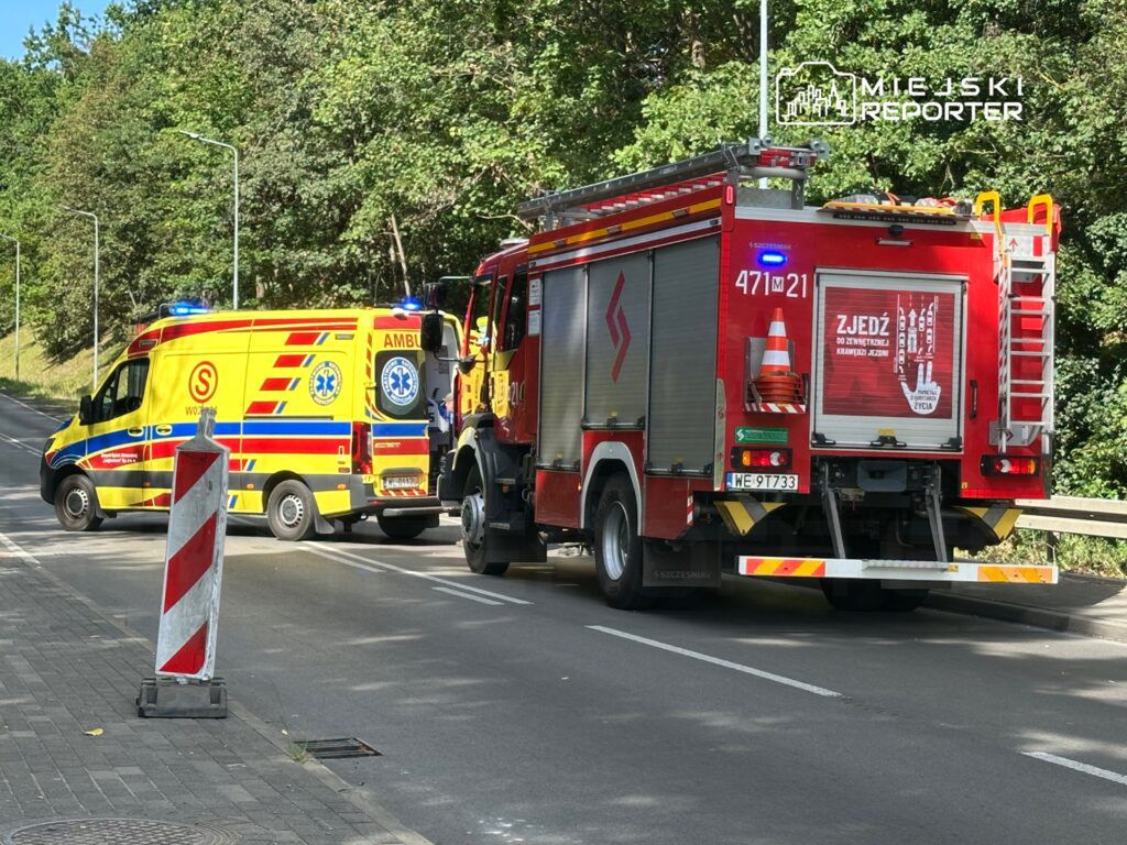 Karetka pogotowia i straż pożarna zatrzymane na drodze, obok znak ostrzegawczy o robót drogowych, w otoczeniu zieleni.