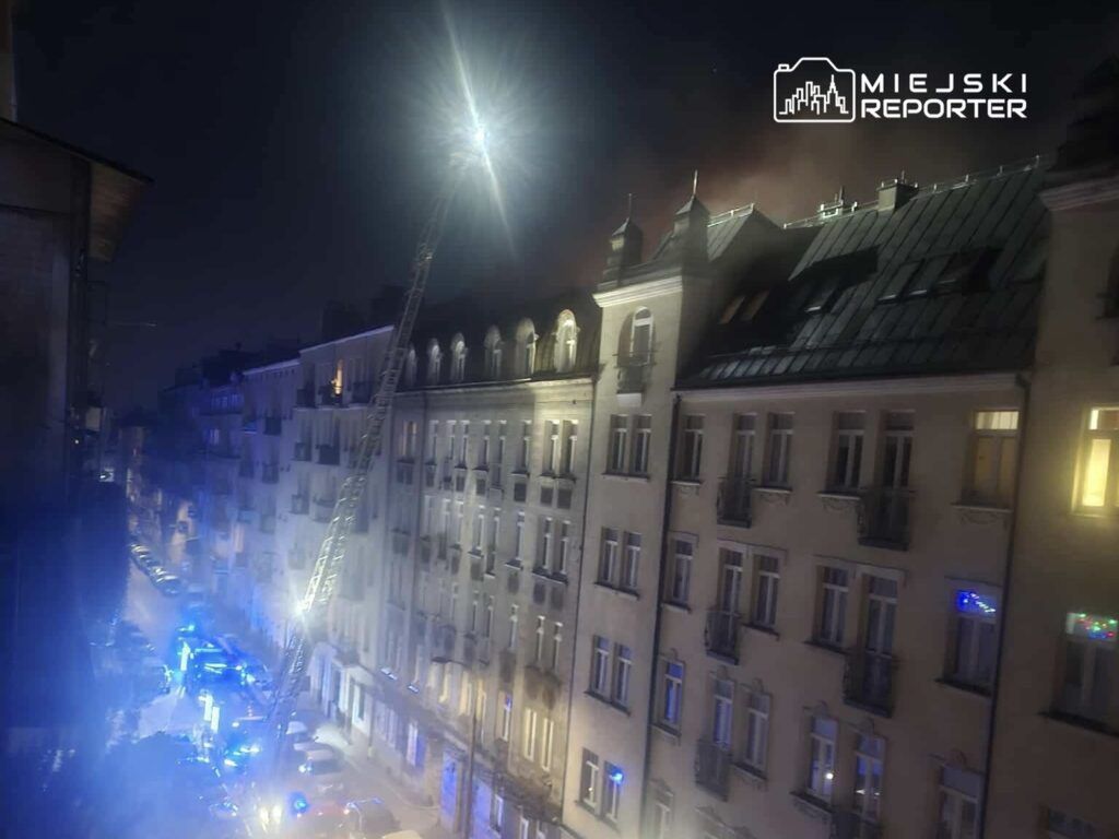 W nocy strażacy używają podnośnika do gaszenia pożaru na dachu budynku w miejskiej okolicy, otoczeni niebieskimi światłami.
