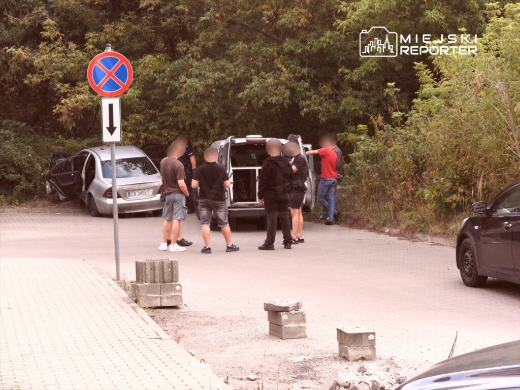 Grupa mężczyzn rozmawia obok otwartego bagażnika furgonetki w pobliżu zaparkowanego samochodu osobowego.