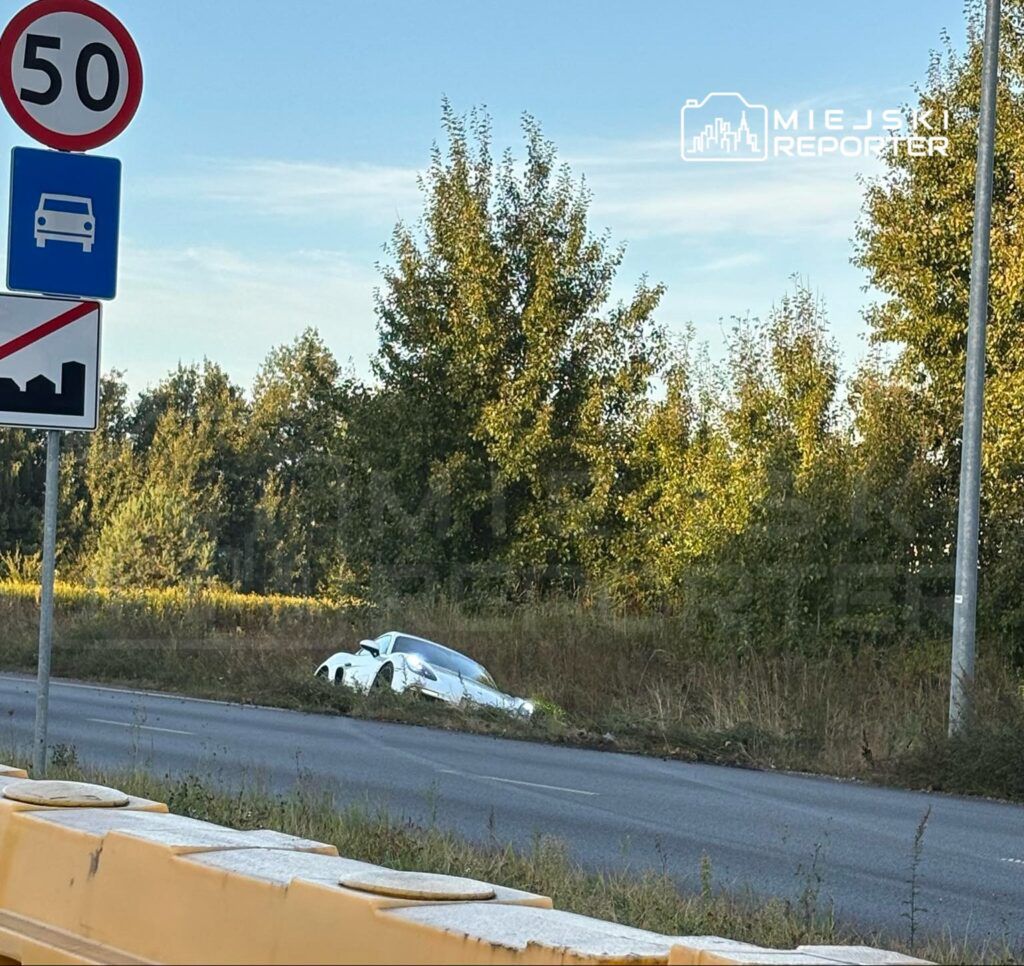 Biały samochód osobowy utknął w zaroślach przy drodze, obok znaków drogowych z ograniczeniem prędkości.