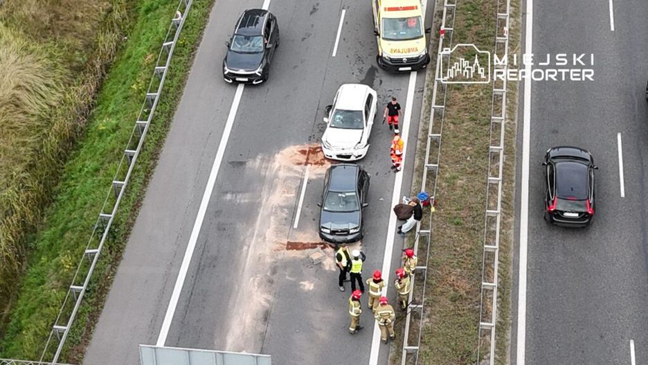 Na drodze doszło do kolizji dwóch samochodów; obok pracują strażacy i ratownicy medyczni.