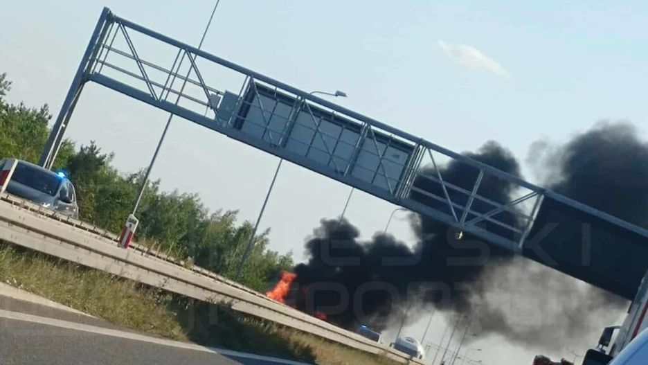 Pożar pojazdu na poboczu drogi, z dymem unoszącym się w powietrzu, obok policyjny radiowóz.