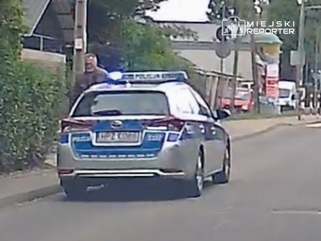 Mężczyzna w brązowej kurtce rozmawia z policjantem obok radiowozu na miejskiej ulicy.