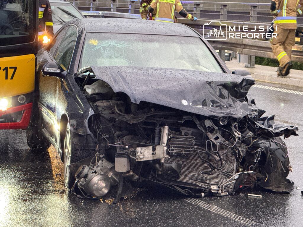 Uszkodzony samochód osobowy z zniszczonym przodem stoi na mokrej jezdni, obok pracują strażacy.