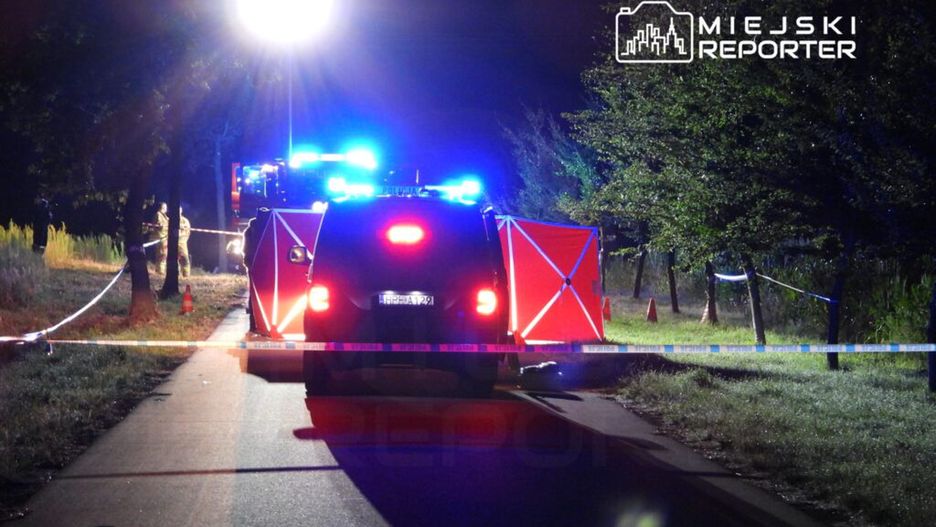 Policyjna akcja w nocy na drodze, z widocznym radiowozem i światłami sygnalizacyjnymi. Funkcjonariusze zabezpieczają teren, na którym rozstawione są taśmy ostrzegawcze. W tle widać pojazdy służb ratunkowych. Miejsce zdarzenia budzi poważne zainteresowanie lokalnych mediów.