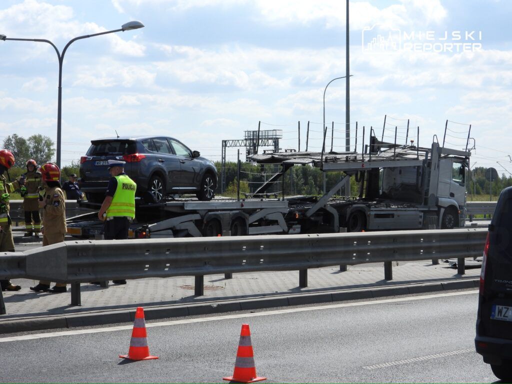 Grupa strażaków i policjantów prowadzi działania na drodze, obok ciężarówki transportującej samochód osobowy.