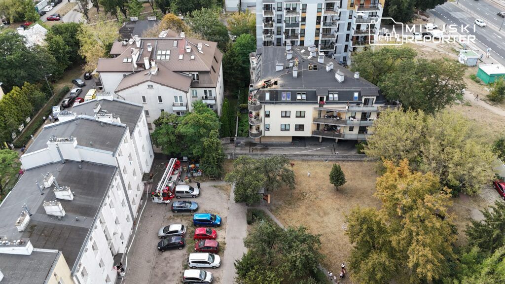 Na dachu budynku mieszkalnego pracują strażacy, obok stoi wóz strażacki, a w tle widoczne są inne budynki i zieleń.