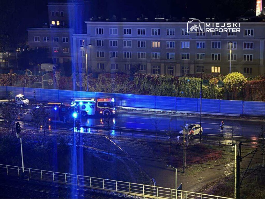 W nocy, na oświetlonej ulicy, straż pożarna oraz policja zabezpieczają miejsce wypadku drogowego.