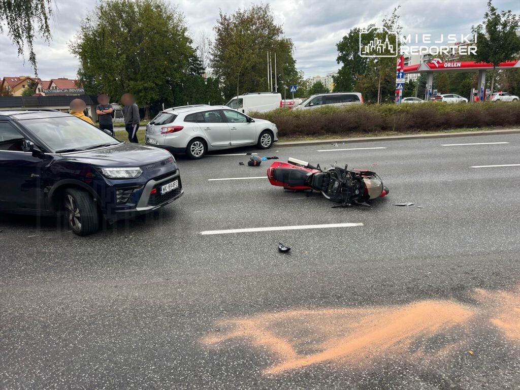 Motocykl leżący na jezdni obok dwóch samochodów, obok grupy ludzi rozmawiających w tle.