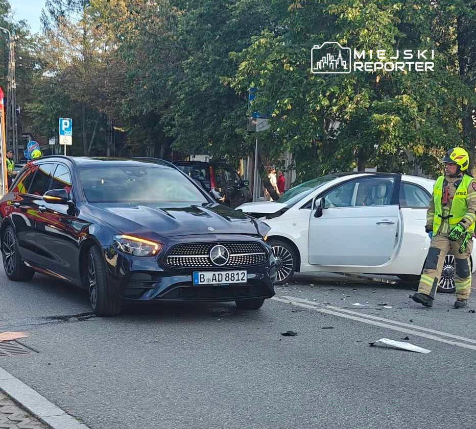 Czarny samochód osobowy z przodu, obok uszkodzony biały pojazd, a obok nich strażak w mundurze, zbierający szczątki.