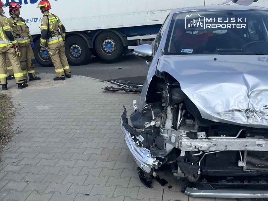 W pobliżu uszkodzonego samochodu osobowego stoją strażacy w mundurach, analizując sytuację po wypadku drogowym.