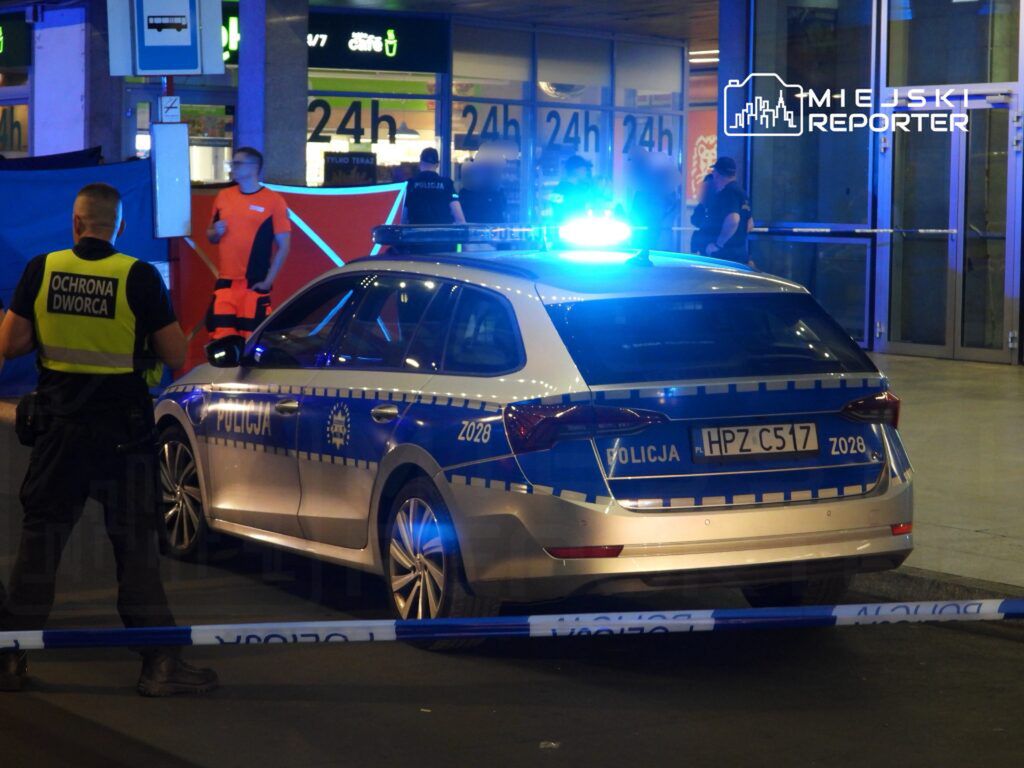 Policjant w kamizelce ochrony dworca obserwuje policyjny samochód z włączonymi sygnałami świetlnymi.