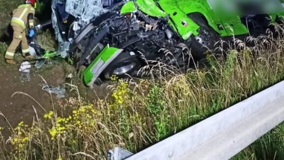 Zielony autobus przewrócony na poboczu drogi, obok strażak w mundurze przeszukuje wrak w nocy.