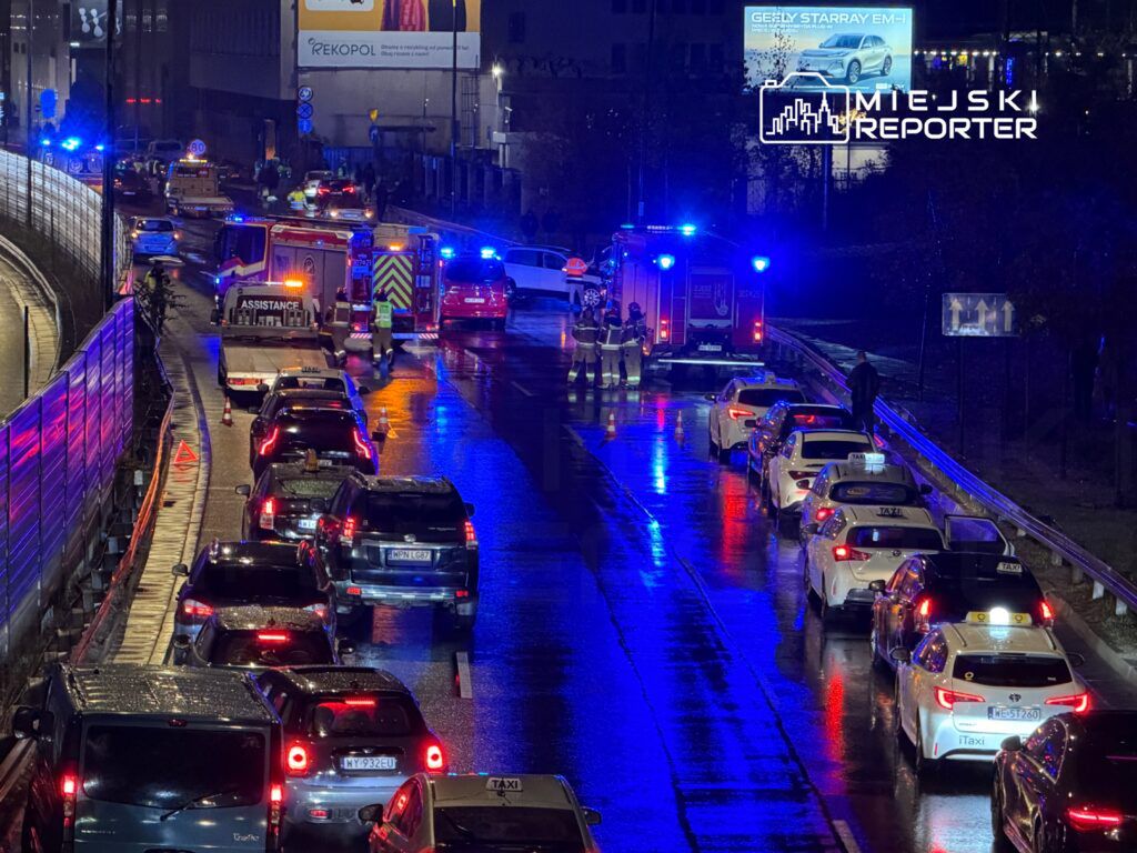 Karetki pogotowia i strażacy pracują na mokrej, oświetlonej niebieskimi światłami drodze, gdzie utknęły samochody.