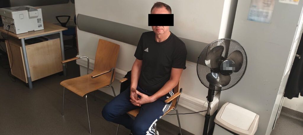 Mężczyzna w czarnej koszulce siedzący na krześle w pomieszczeniu z biurkiem i wentylatorem.