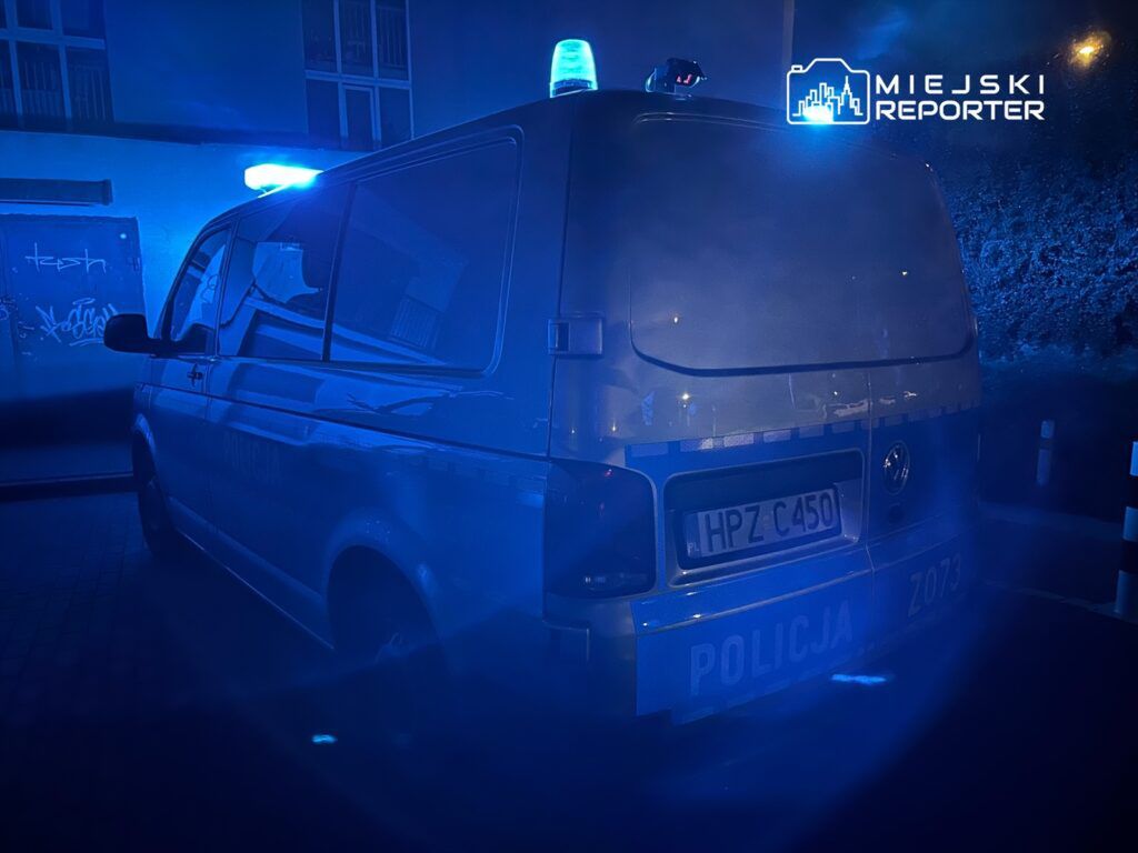 Policyjny radiowóz z niebieskimi światłami stojący w ciemnej uliczce, w tle graffiti na ścianie.