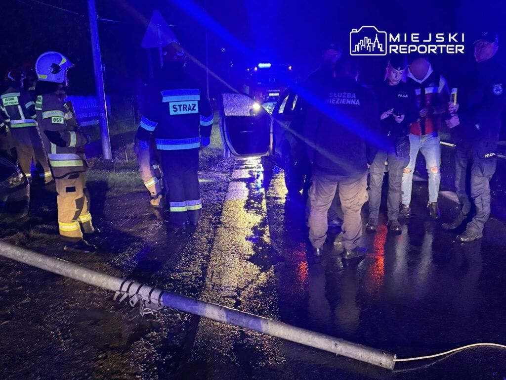 Grupa strażaków i policjantów zbiera się wokół uszkodzonego pojazdu na mokrej drodze, obok leżącego słupa.