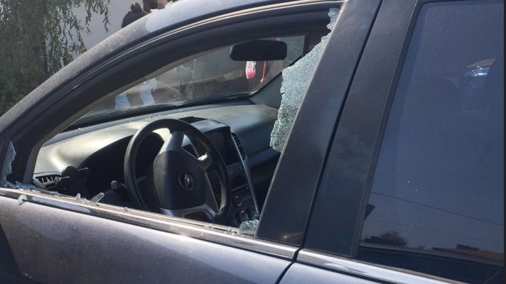 Uszkodzone auto z wybitym oknem, w środku widoczna kierownica, w tle osoby stojące obok budynku.